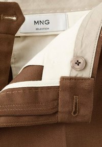 Tessuto marrone di un indumento con un accento beige e dettagli di bottone e zip; etichetta MNG visibile, che indica il marchio e la selezione.