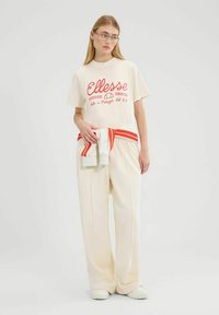 Camiseta color crema con estampado rojo de "Ellesse", combinada con pantalones fluidos color crema. Ceñida con una tela de rayas blancas y rojas.