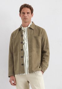 Olivgrüne Wildlederjacke mit Knopffront, zwei Brusttaschen und Kragen, kombiniert mit einem weißen Hemd mit grünen Streifen.