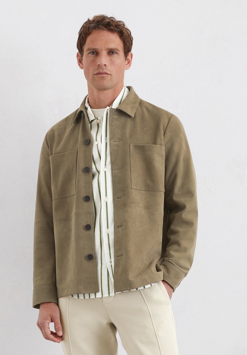 Olivgrüne Wildlederjacke mit Knopffront, zwei Brusttaschen und Kragen, kombiniert mit einem weißen Hemd mit grünen Streifen.