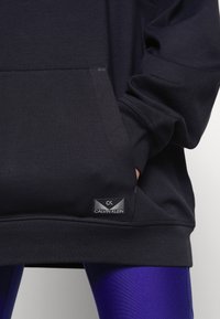 Felpa nera realizzata in morbido materiale, con tasca frontale e etichetta Calvin Klein all'orlo. Abbinata a leggings viola lucidi.
