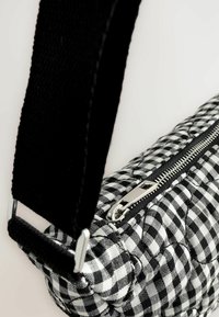 Gepäcksbeutel im gesteppten schwarz-weißen Schachbrettmuster mit einem silbernen Reißverschluss und einem schwarzen Stoffriemen. Verfügt über eine gestickte Textur und eine abgerundete Form.