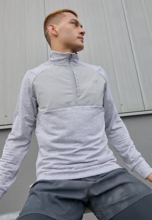 Lys grå atletisk genser med kvart glidelås, teksturert stoff og en ensfarget frontpanel. Brukt med mørke shorts, mot en grå bakgrunn.