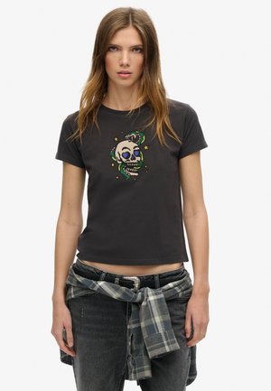 Camiseta de algodón negra con mangas cortas, que presenta un diseño colorido de una calavera rodeada de estrellas y una serpiente. Se lleva con una camisa de cuadros anudada a la cintura.