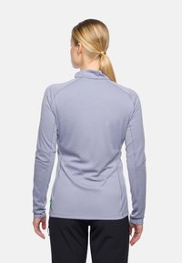 Eine Frau mit einem blonden Pferdeschwanz trägt ein leicht lila langärmliges Oberteil und schwarze Hosen und steht mit dem Rücken zu einem schlichten weißen Hintergrund.