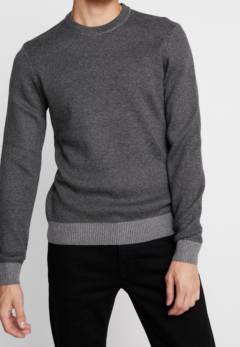 Grå stickad tröja med fiskbensmönster, rund halsringning och ribbade muddar och fåll. Matchas med svarta byxor.