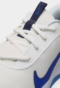 Weißer Sportschuh mit weißen Schnürsenkeln, strukturiertem Obermaterial und blauem Nike-Swoosh-Logo an der Seite sowie am Schnürsenkel-Ösenband.