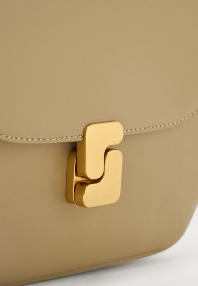 Μπεζ δερμάτινη τσάντα με λεπτομερή κοντινή λήψη του γυαλισμένου χρυσού μεταλλικού κούμπωμα και ορατή ραφή στην άκρη της παλάμης.