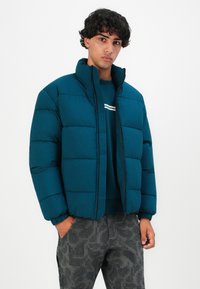 Joven vestido con una chaqueta tipo plumón verde azulado sobre una sudadera azul con una francha blanca, pantalones de camuflaje digital gris, de pie con una mano en el bolsillo.