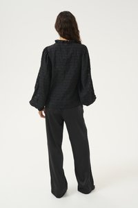 Black textured blouse with oversized puff sleeves and a ruffled collar, paired with loose-fitting black trousers. Minimalistic design.
