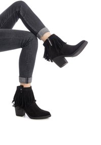 Stivaletti neri in suede alla caviglia con dettagli in frange, una cerniera laterale e un tacco a blocco testurizzato, abbinati a jeans scuri risvoltati.