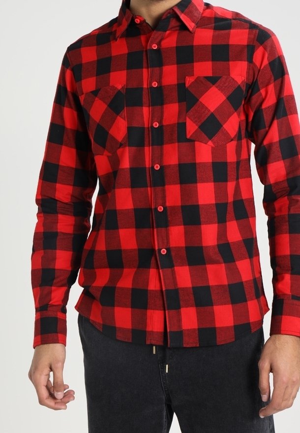 Homme portant une chemise en flanelle à carreaux rouges et noirs, boutonnée, avec deux poches poitrine et un pantalon noir, faisant face à un fond uni.