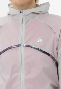 Chaqueta ligera con cremallera en rosa suave y capucha. Presenta detalles plateados y un diseño texturizado sutil. Logo exhibido en el pecho.
