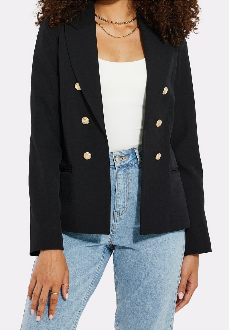 Blazer noir croisé à double boutonnage avec des boutons dorés, au design structuré et avec des revers crantés. Porté sur un haut blanc, associé à un jean bleu clair.