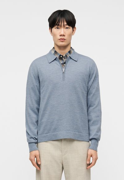 Homme portant un pull à demi-fermeture éclair bleu clair par-dessus une chemise à carreaux bleus et beiges avec un pantalon beige, debout devant un fond blanc uni.