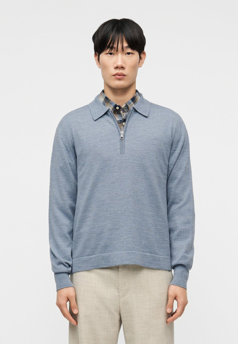 Άνδρας που φοράει ελαφρύ πουλόβερ  με φερμουάρ μέχρι το μέσο του στήθους σε ανοιχτό μπλε χρώμα πάνω από μπλε και μπεζ πουκάμισο με καρό σχέδιο, και μπεζ παντελόνι, στέκεται μπροστά σε απλό λευκό φόντο.