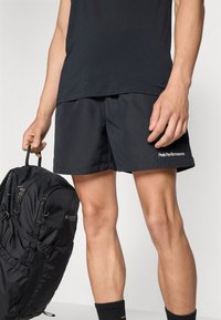 Shorts negri de sport cu o textură netedă, având un logo alb "PeakPerformance". Purtați cu un rucsac negru și ciorapi până la gleznă.