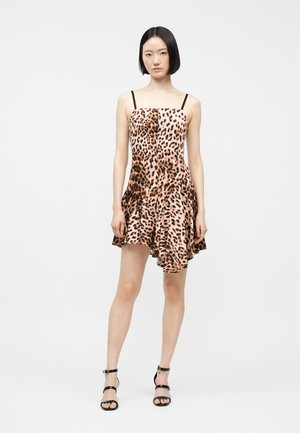 Žena v minišatech bez rukávů s tygřím vzorem, černými ramínky a černými sandály na vysokém podpatku, stojící na jednobarevném pozadí.