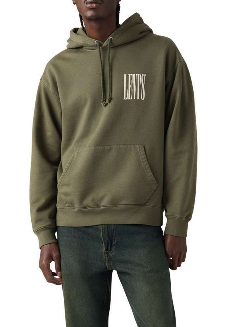 Hombre con sudadera con capucha Levi's de color verde oliva con bolsillo delantero y cordones, combinado con vaqueros azules, de pie frente a un fondo blanco.