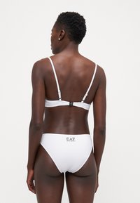 Biely bikiny set s minimalistickým dizajnom, tenkými popruhmi a logom na zadnej strane spodného dielu. Hladká textúra a priliehavý strih.