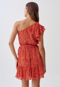 Robe rouge à fleurs, à une épaule, avec une manche à volants, une taille froncée et une jupe à volants. Tissu léger et transparent avec une texture fluide.