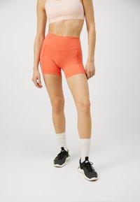 Shorts de sport orange taille haute en tissu extensible, associés à des chaussures de sport noires et des chaussettes blanches mi-mollet. Design minimaliste.