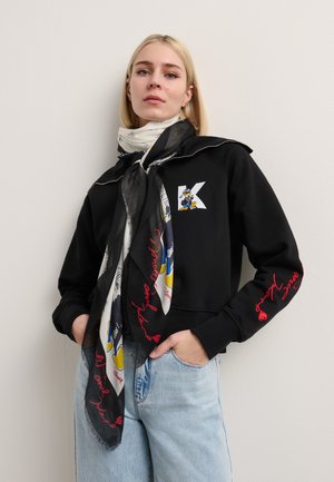 Schwarzer Pullover mit einem weißen "K"-Logo, kombiniert mit einem gemusterten Schal mit Cartoonfiguren und rotem Text, getragen mit hellblauen Jeans.