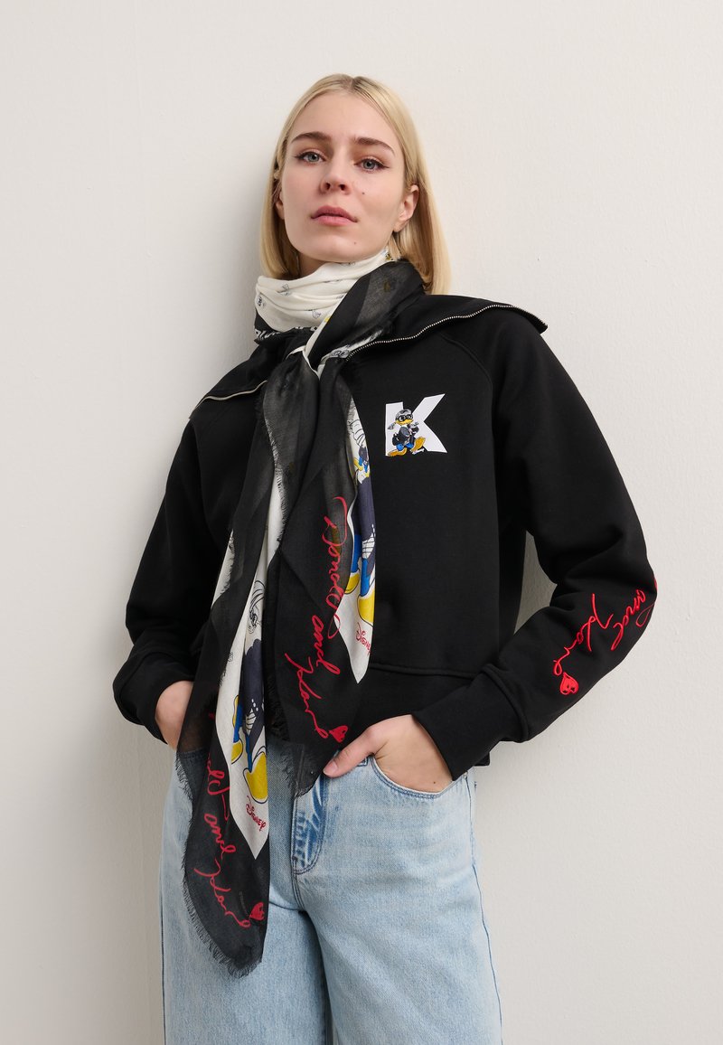 Svart sweatshirt med en hvit "K"-logo, kombinert med et mønstret skjerf som har tegneseriefigurer og rød tekst, brukt sammen med lyseblå jeans.