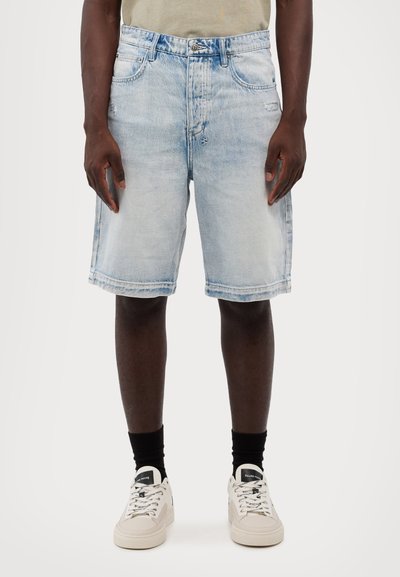 Calções de ganga azul claro com um corte descontraído, apresentam cinco bolsos, fecho com botão e uma bainha desfiada. Combinados com ténis brancos e meias pretas.