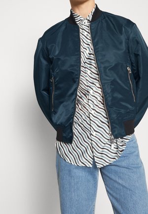 Homme portant une veste bomber bleu foncé par-dessus une chemise blanche à rayures ondulées noires et bleues, associée à un jean bleu clair, mains dans le dos.