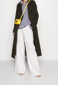 Olivgrön lång kappa, marinblå och vit randig polo, vita vida byxor och en liten gul crossbody-väska. Sandaler med remmar kompletterar looken.