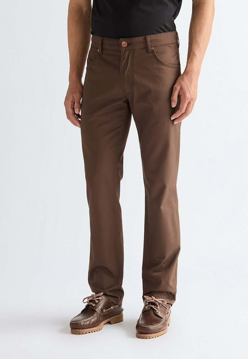 Homme portant un pantalon droit marron et des chaussures en cuir marron foncé à lacets, debout devant un fond clair uni.