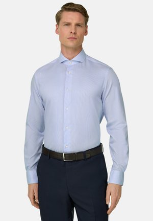 Mężczyzna w jasnoniebieskiej koszuli z długim rękawem wpuszczonej w ciemne spodnie z czarnym paskiem, stojący na jednolitym tle.