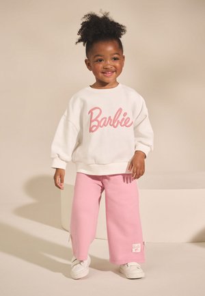 Usměvavé batole s kudrnatými vlasy v bílém mikině "Barbie", růžových kalhotách a bílých teniskách stojící na neutrálním pozadí.