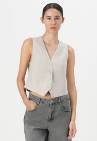 Gilet beige cropped con righe verticali, scollatura a V, quattro bottoni metallici e una vestibilità aderente, abbinato a jeans grigi a vita alta.