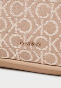 Beżowa torba tekstylna z wytłaczanym wzorem logo. Gładka, skórzana podstawa o subtelnym połysku. Nazwa marki wyświetlona w metalicznym złocie.