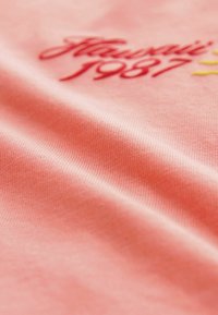 Tela de algodón en color coral con superficie texturizada y diseño bordado que incluye "Hawái 1987" en acentos rojos y amarillos.