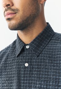 Camicia a quadri abbottonata in tessuto grigio scuro e nero con trama strutturata, caratterizzata da colletto appuntito e bottoni bianchi.