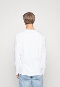 Camiseta blanca de manga larga con cuello redondo, fabricada en algodón. Mostrada desde detrás, combinada con jeans de mezclilla azul claro.