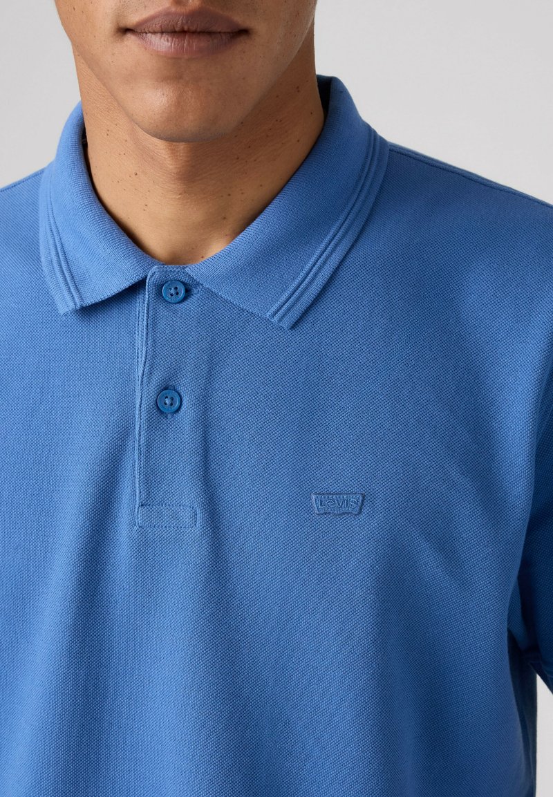 Hombre que lleva una camisa polo azul de Levi's con dos botones abrochados, mostrando el cuello y la parte superior del pecho.