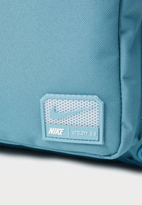 Nike Utility 2.0 laukku vaaleansinisessä kankaassa, jossa on rakenteellinen pinta ja suorakulmainen etiketti, jossa on rei'itetty kuvio ja logo.