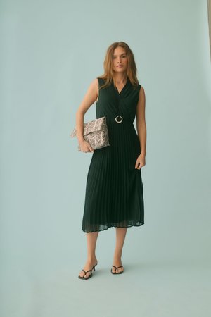 Nő ujjatlan fekete, pliszírozott midi ruhában, kezében nagy kígyóbőr mintás kis táska, fekete, magas sarkú szandált visel, halványkék háttér előtt áll.