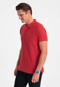 Chemise polo rouge en coton, avec un col classique et des manches courtes. Portée avec un jean en denim foncé et une montre bleue.
