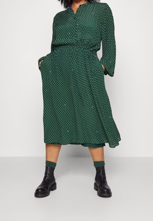 Kobieta w zielonej sukience midi z długim rękawem w grochy z kieszeniami, zielonych skarpetkach i czarnych masywnych botkach, stojąca z rękami w kieszeniach.