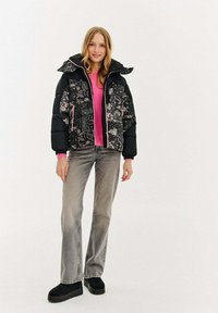Kobieta stojąca w czarnej pikowanej kurtce w kwiatowy wzór, różowym swetrze, szarych dżinsach i czarnych butach na platformie na białym tle.
