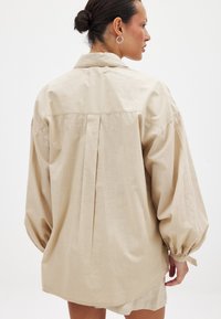 Beige oversized skjorta med en ryggveck, mjuk textur, långa fladdrande ärmar och knapppdetaljer vid ärmsluten. Slät tygfinish.