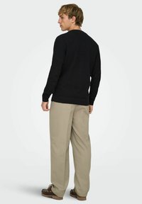 Mustast kudumist kampsun pikkade varrukatega, ümar kaelusega ja soonikkoes äärega, kombineeritud beežide volditud pükste ja pruunide kingadega. Tagantpoolt vaade.