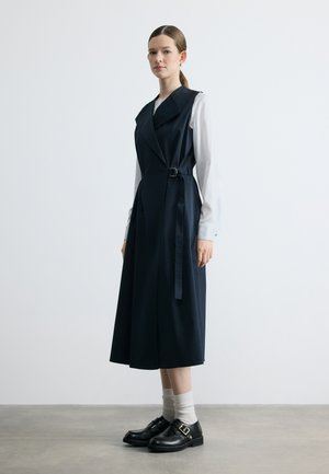 Femeie stând lângă un perete alb, purtând o rochie midi bleumarin fără mâneci, cu curea, o cămașă albă cu mâneci lungi, șosete albe și pantofi negri cu cataramă.