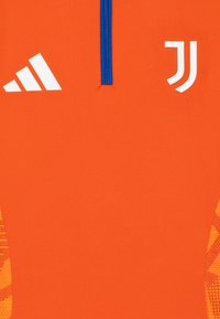Narandžasta atletska majica s plavim vezicama, s bijelim logotipima Adidasa i Juventusa. Teksturalni uzorci u tamnije narančastoj boji na rukavima.