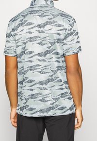 Košile s krátkým rukávem ve světle šedé barvě s tmavě modrým a černým vlnitým vzorem, která má stojáček a hladkou, strukturovanou tkaninu.